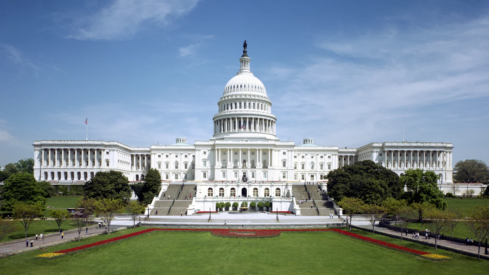 The west front of the U.S. Capitol seen across its lawn under a blue sky.