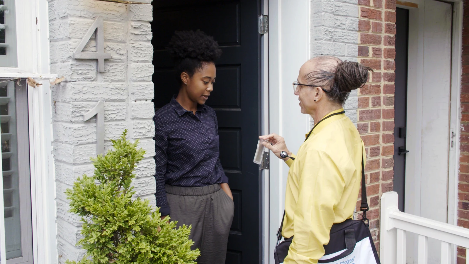 A Census Bureau field worker wearing a yellow shirt speaks with a resident at the doorway of a brick home.