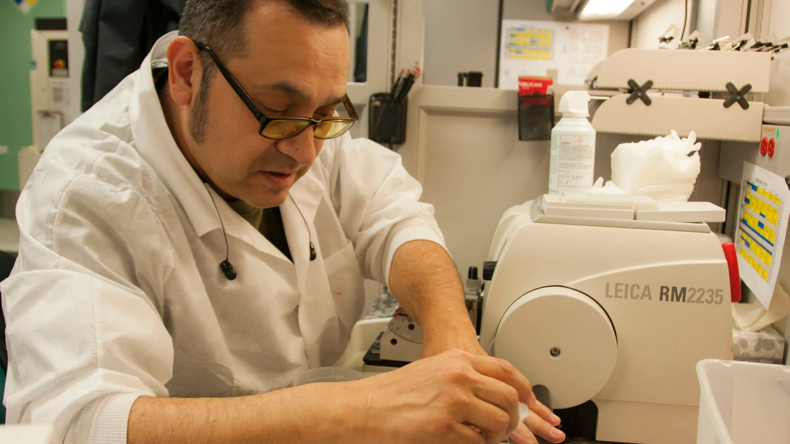 A histology technician in a white coat prepares a very thin tissue sample beside laboratory equipment.