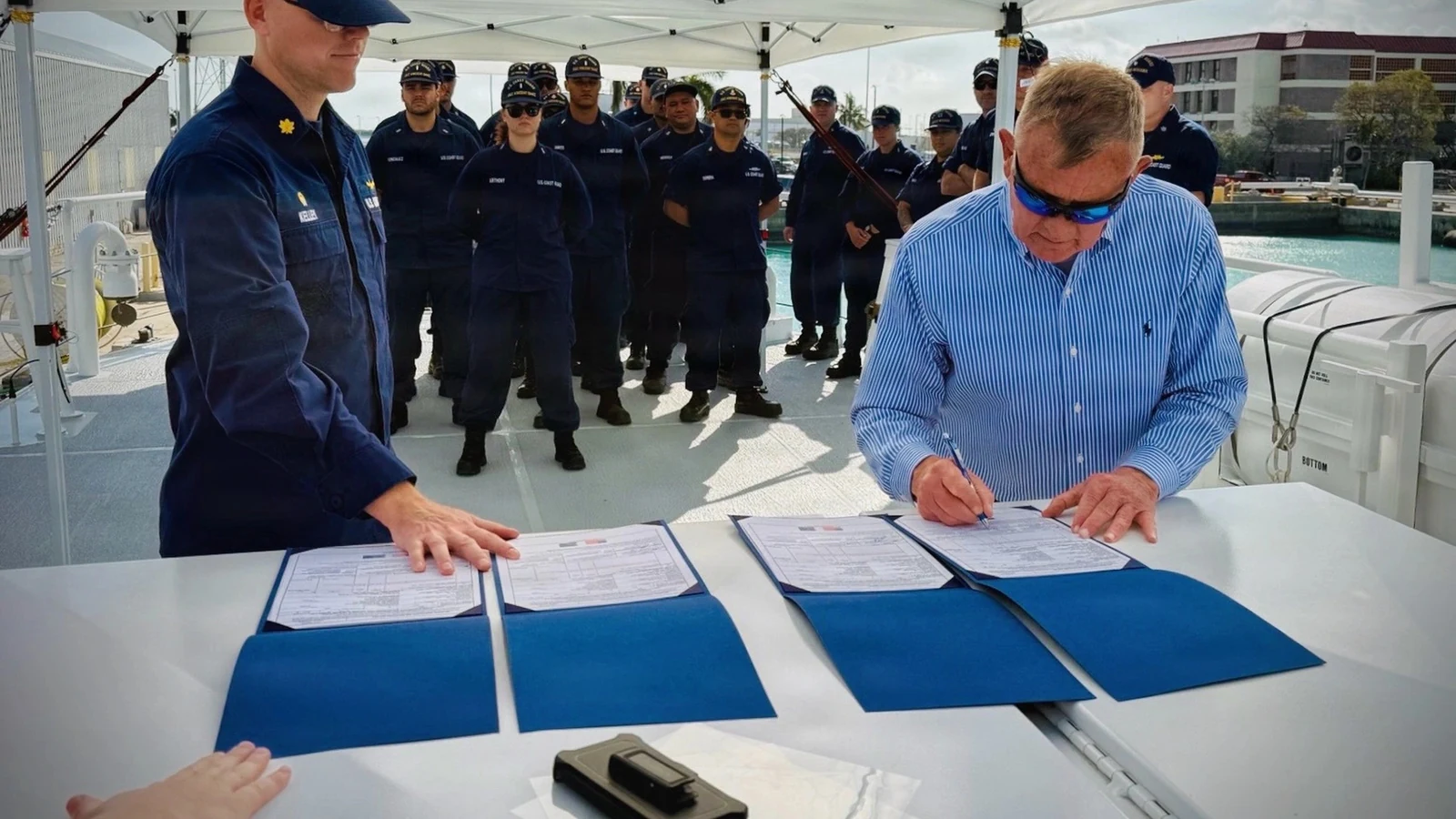 A Coast Guard contracting officer signs acceptance documents on a cutter while uniformed crew members watch.