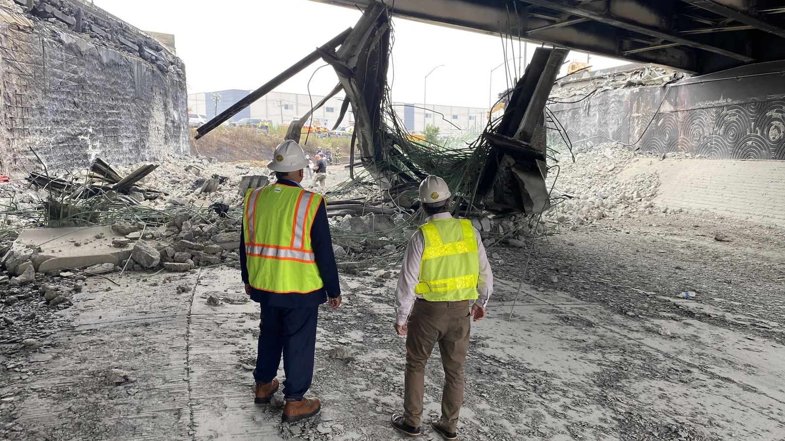 Federal Highway Administration officials in safety gear inspect damaged concrete beneath an elevated highway.