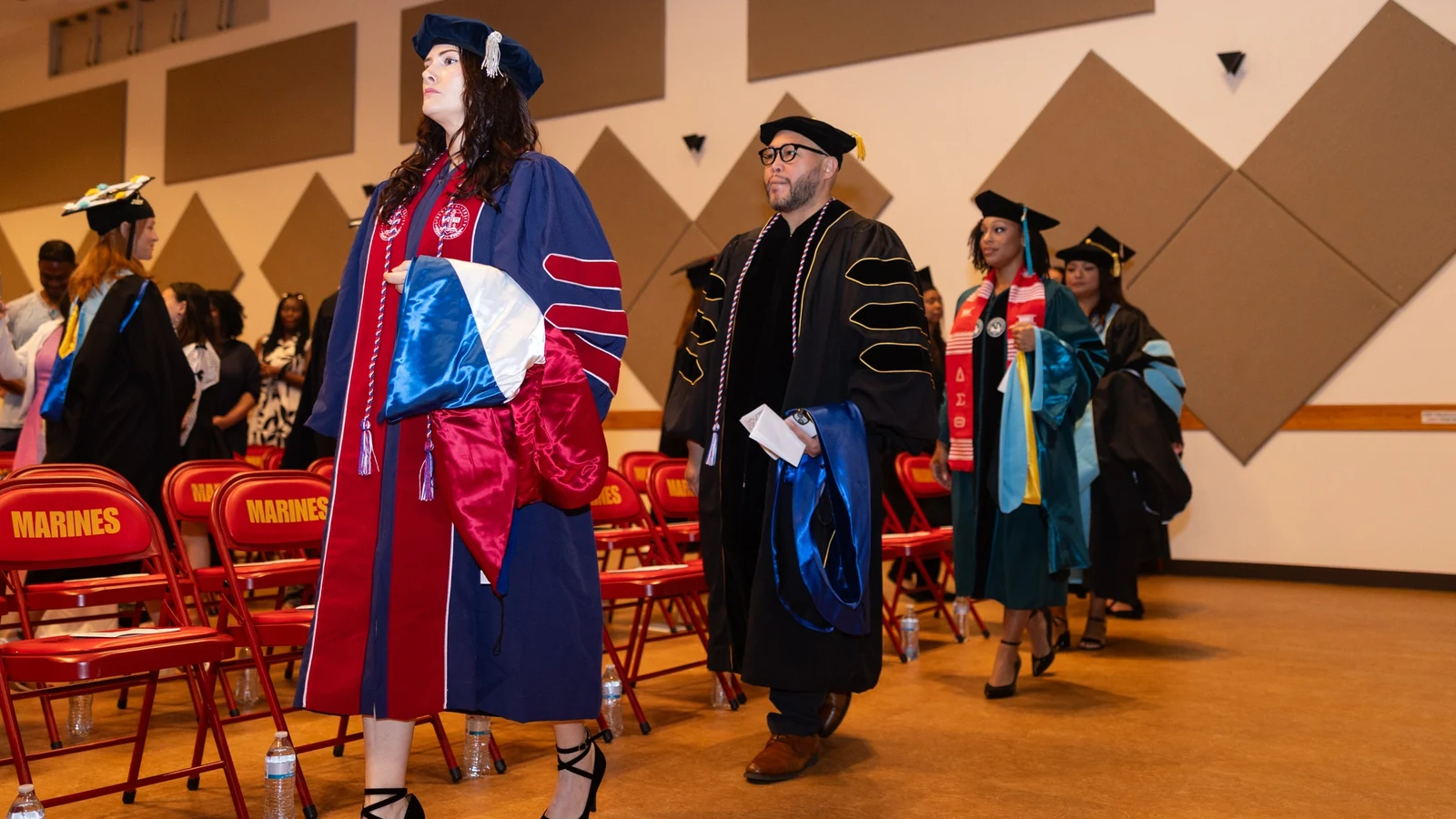 College graduates in caps and gowns stand beside faculty at a postsecondary commencement ceremony.