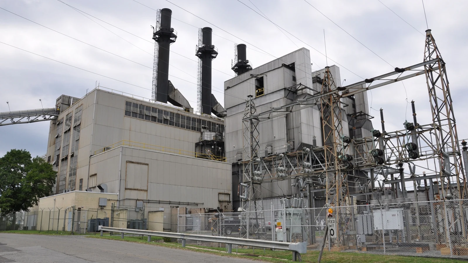 An industrial power plant with three smokestacks stands behind electrical equipment and transmission lines.