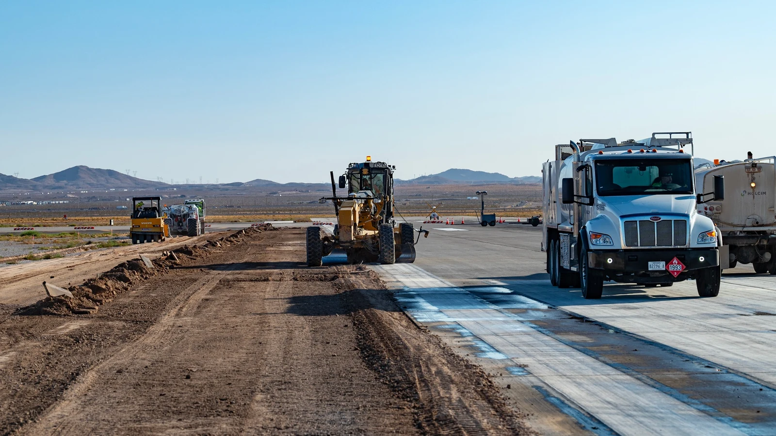 Construction workers and heavy machinery spread material across an airport runway under a wide blue sky.