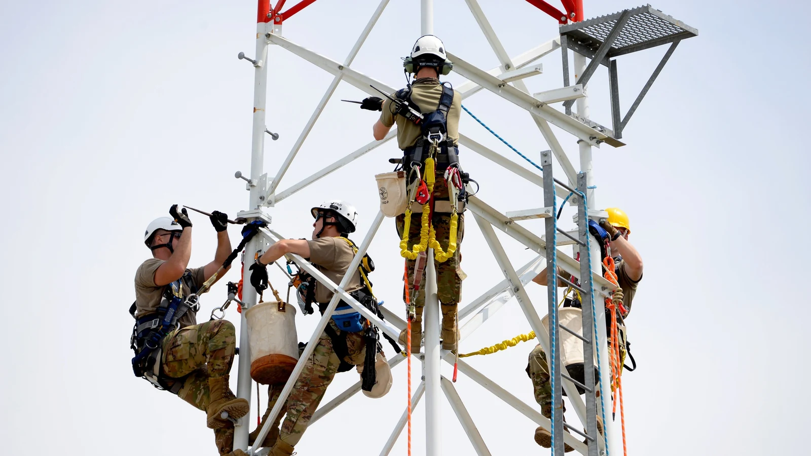 Airmen in safety harnesses assemble a communications tower against a clear sky.
