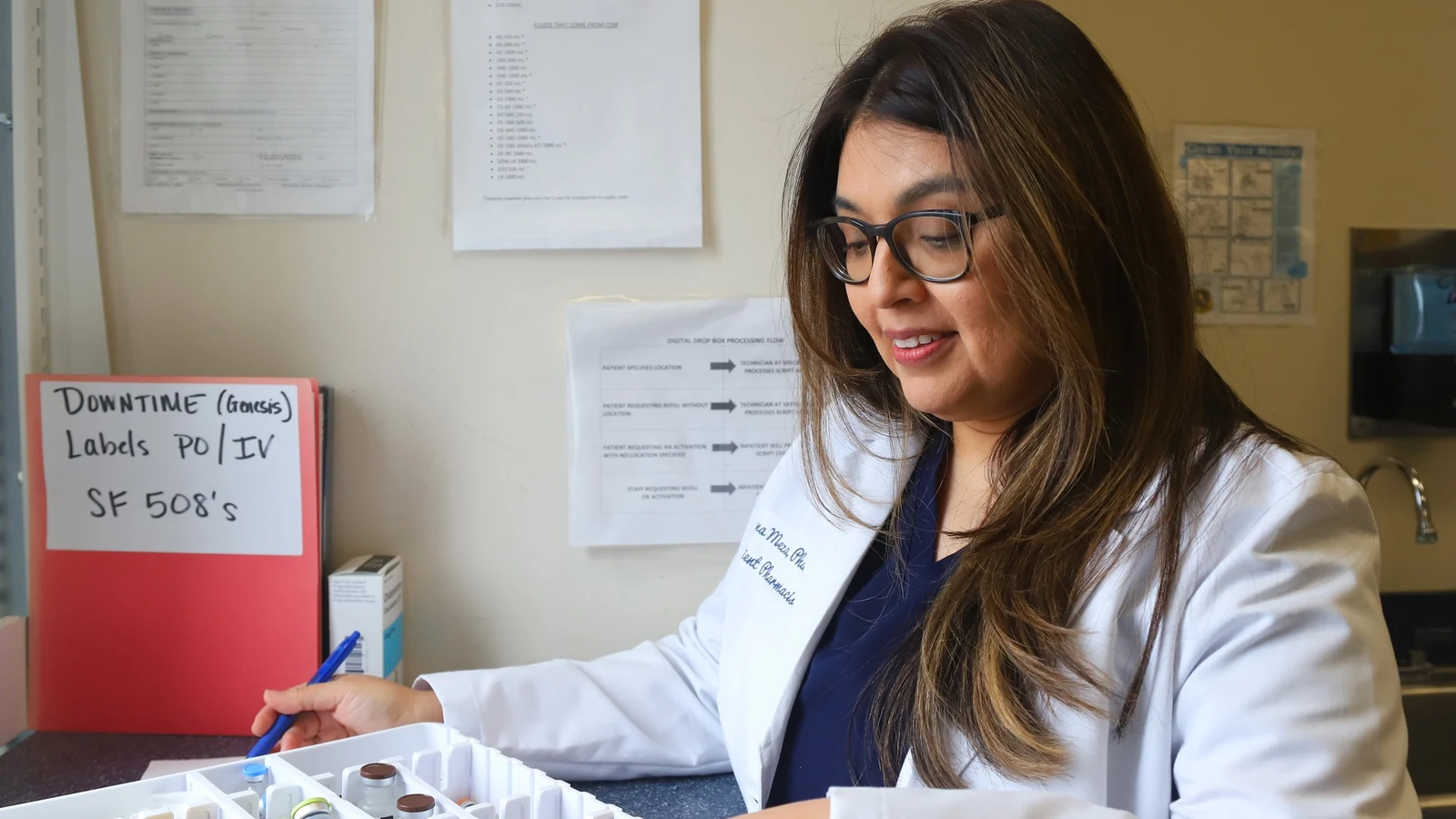A pharmacist in a white coat checks medications in bins at a hospital pharmacy.