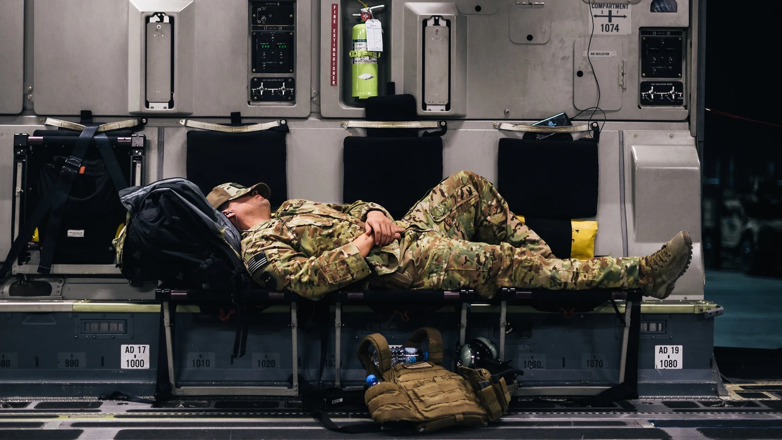 An airman sleeps in a dim aircraft cabin while other personnel sit nearby.
