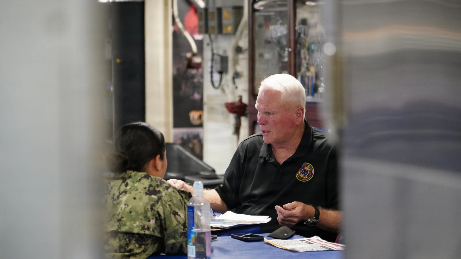 A researcher briefs a sailor while holding a small wearable health device aboard a ship.
