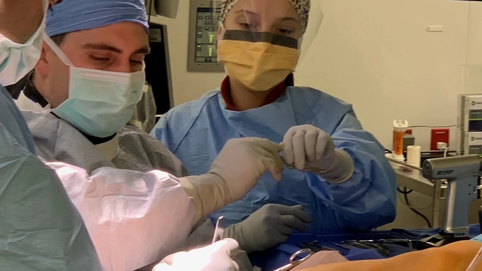 Three masked clinicians perform ankle surgery under bright operating-room lights.