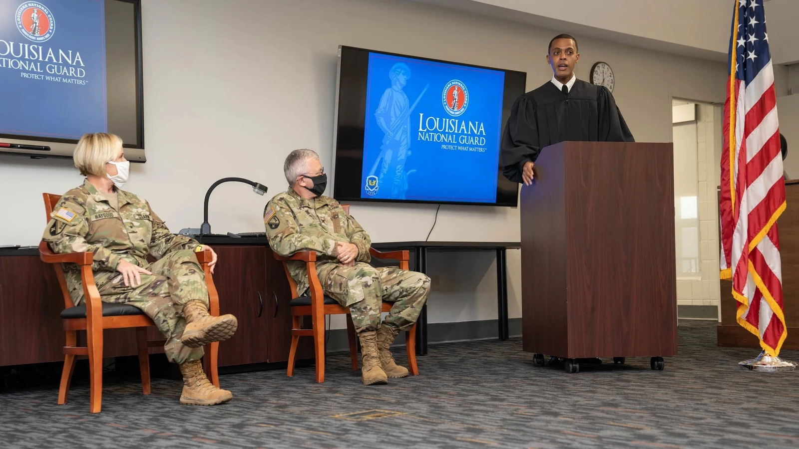 A military judge speaks at a lectern in a courtroom while three uniformed officials sit nearby.
