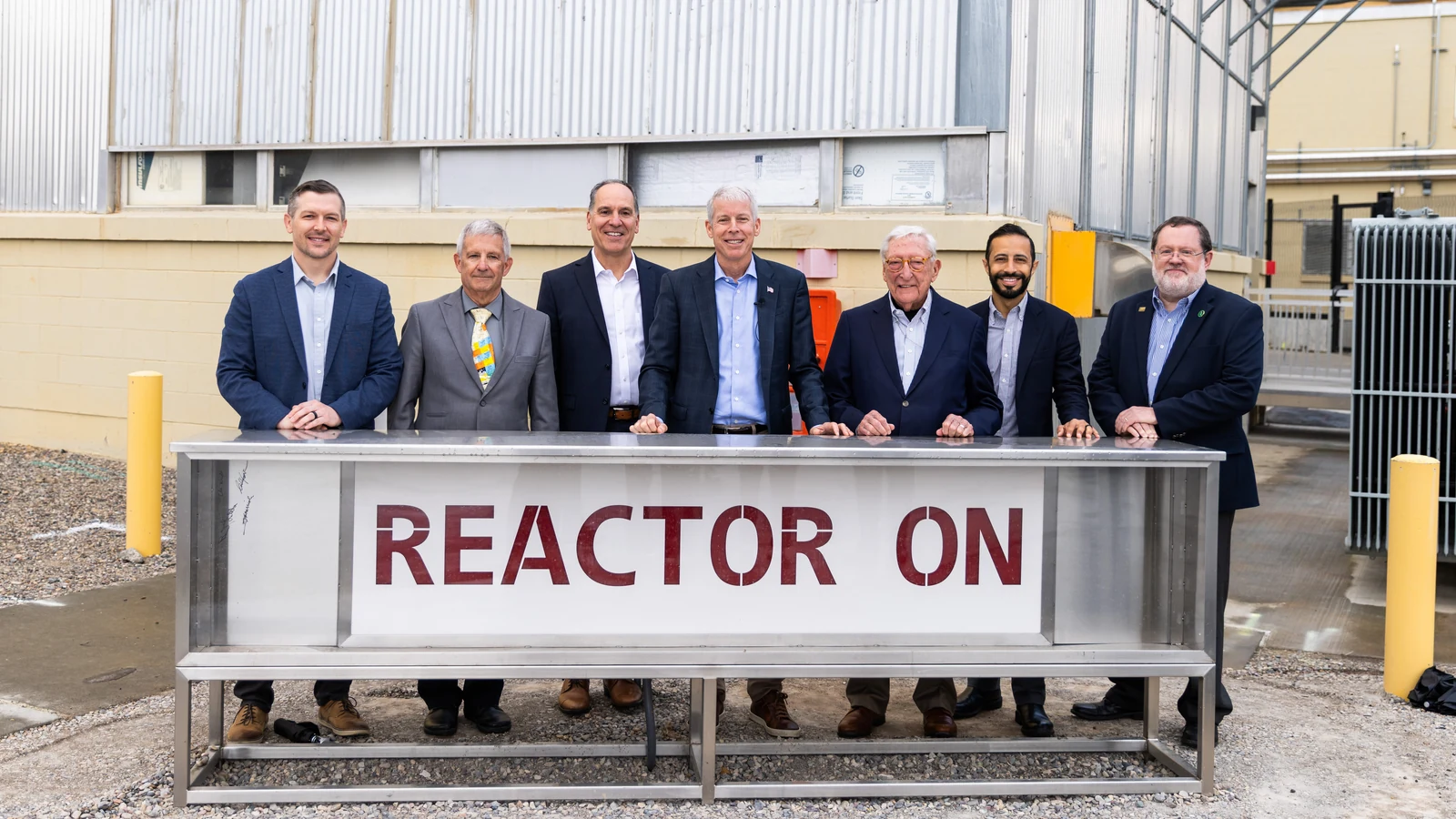 Seven Energy Department and Idaho National Laboratory officials stand behind a large metal sign reading “Reactor On” outside an industrial test building.