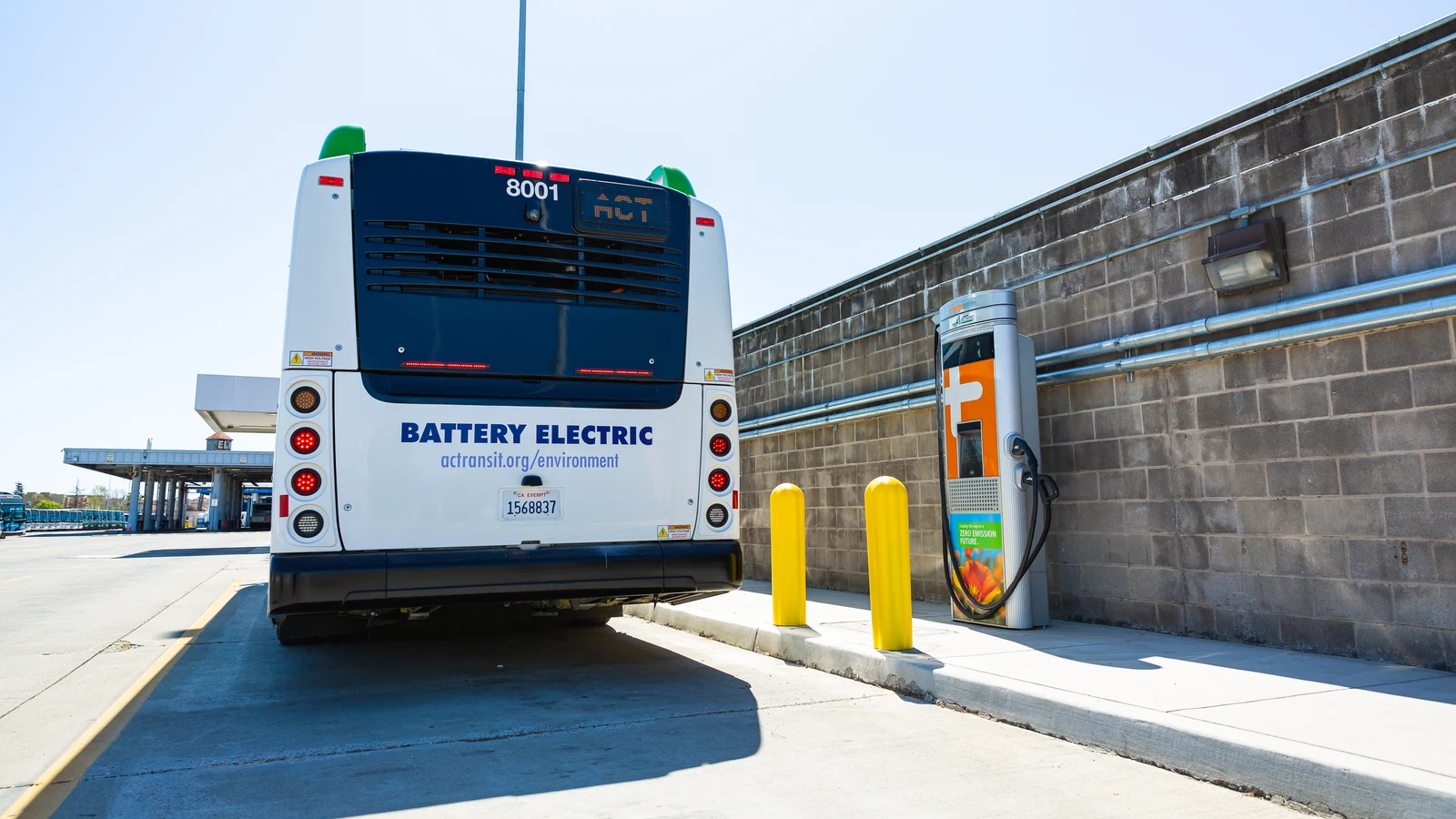 A battery-electric AC Transit bus stands beside a charging station at a California depot.