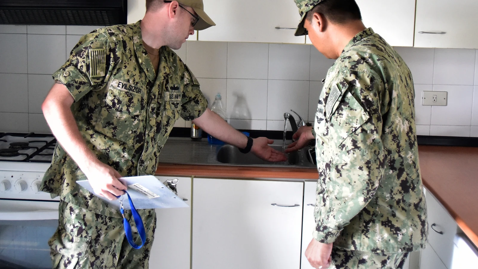 Two housing inspectors examine the sink and fixtures inside an apartment kitchen.