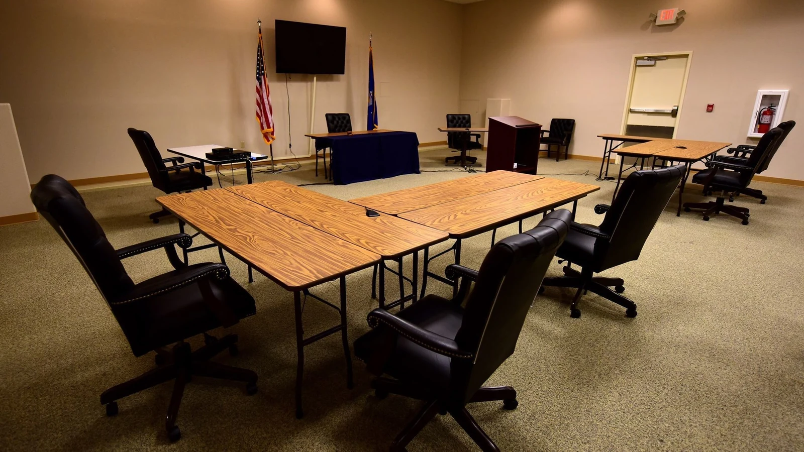 An empty courtroom contains counsel tables, microphones, a lectern, flags and a wall-mounted video screen.