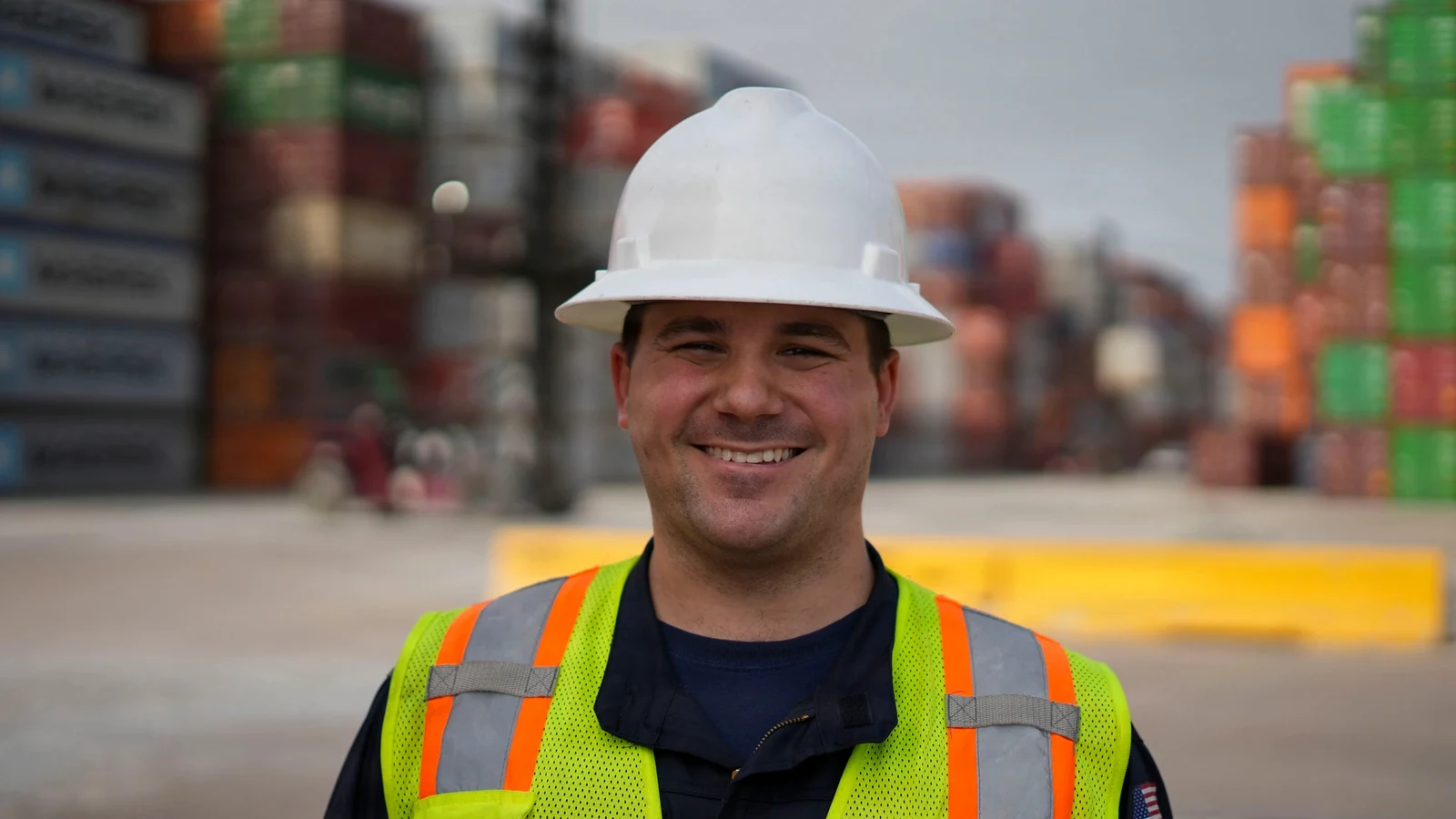 A Coast Guard member in a hard hat and reflective vest stands in front of stacked shipping containers at Port Houston.
