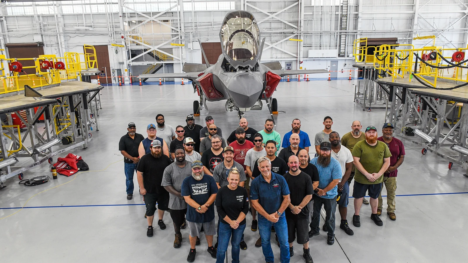 Personnel stand beside an F-35B fighter inside an aircraft maintenance and production hangar.