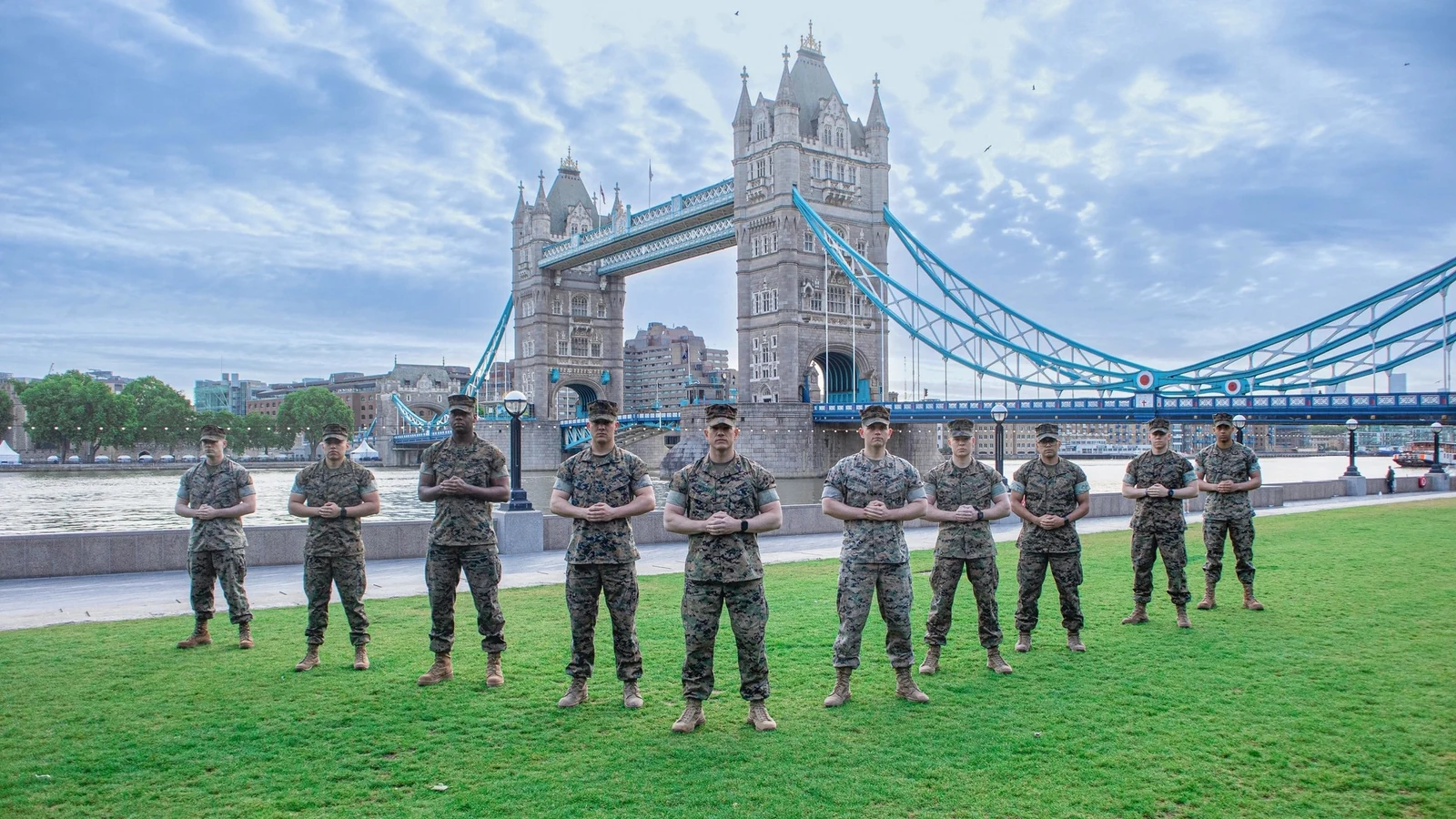 A line of U.S. Marines stands on the Thames embankment with Tower Bridge behind them under a blue sky.