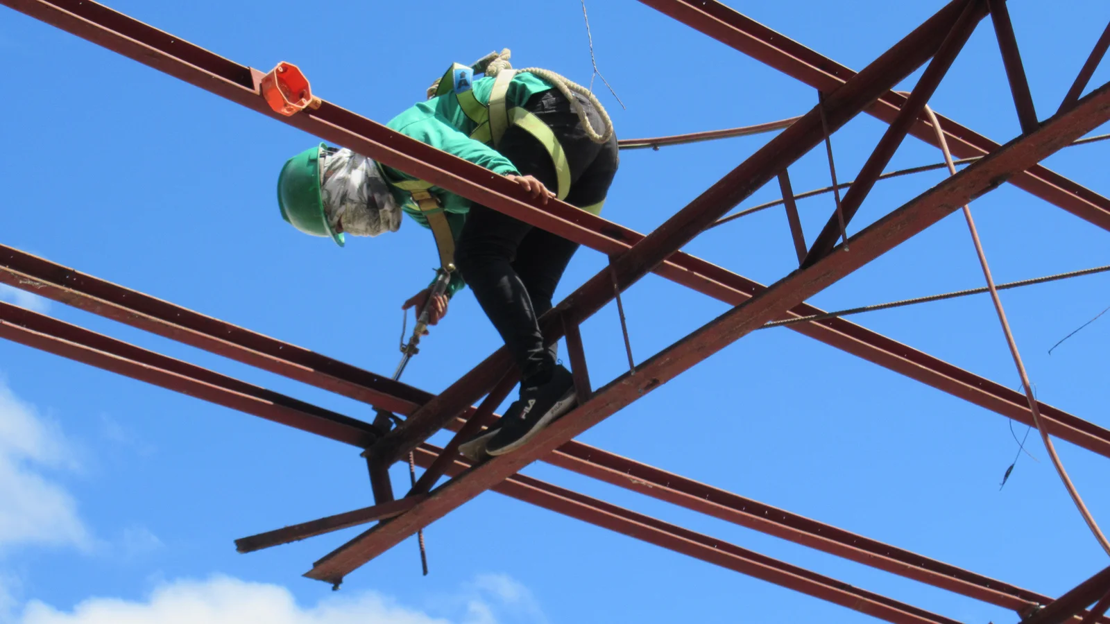 Welder in a green hard hat works high on a metal framework against a blue sky.
