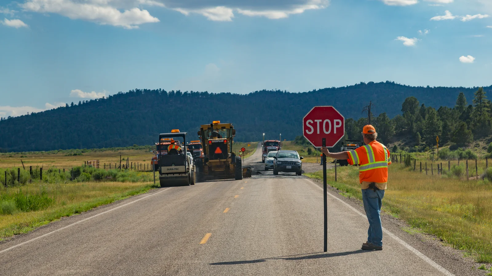 Road construction worker in reflective gear directs traffic beside work vehicles.