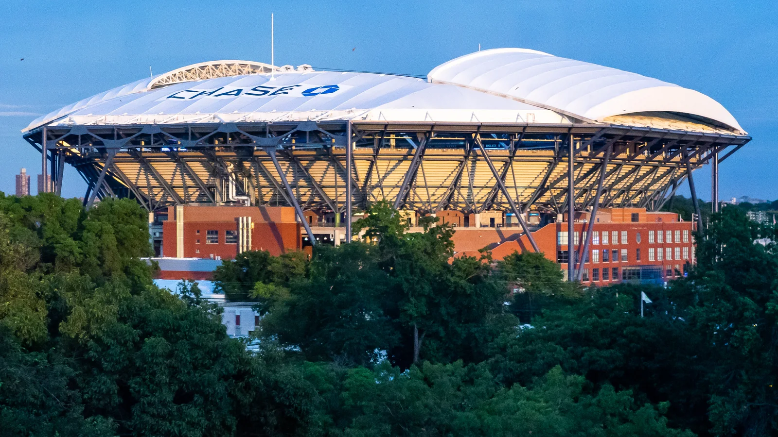 Arthur Ashe Stadium rises above the grounds of the USTA tennis center beneath a blue sky.
