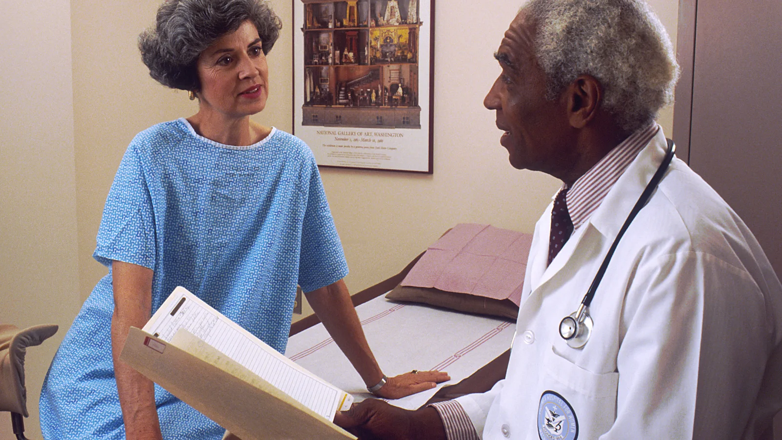 Doctor sits beside a patient during a consultation in a clinic room.