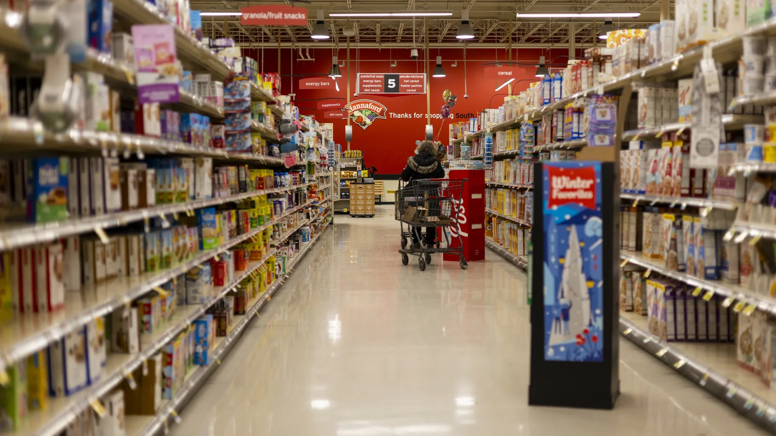 Long supermarket aisle lined with packaged foods under bright ceiling lights.