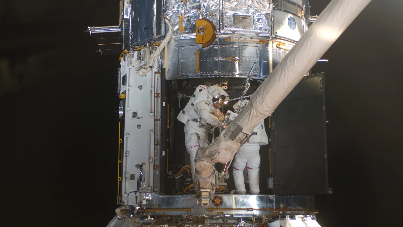 Two astronauts in white spacesuits work on the Hubble Space Telescope above Earth.