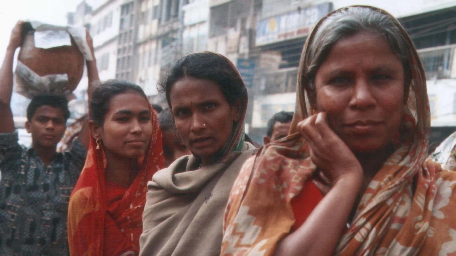 Pedestrians in colorful clothing walk along a busy street in Dhaka.
