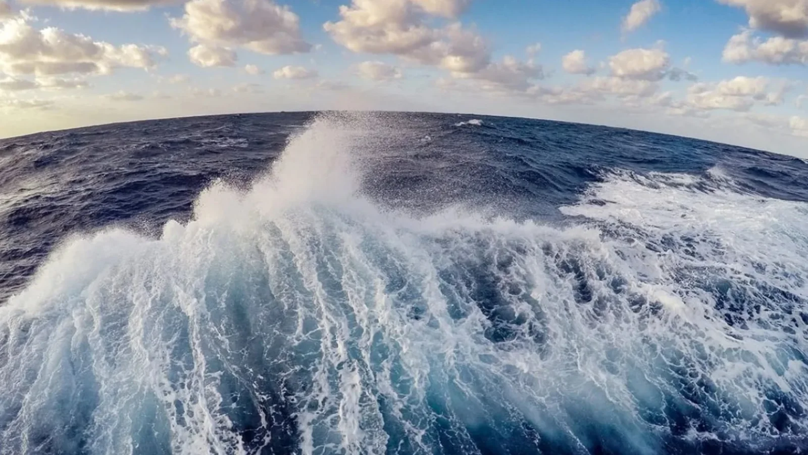 White ship wake stretches across deep blue ocean beneath scattered clouds.