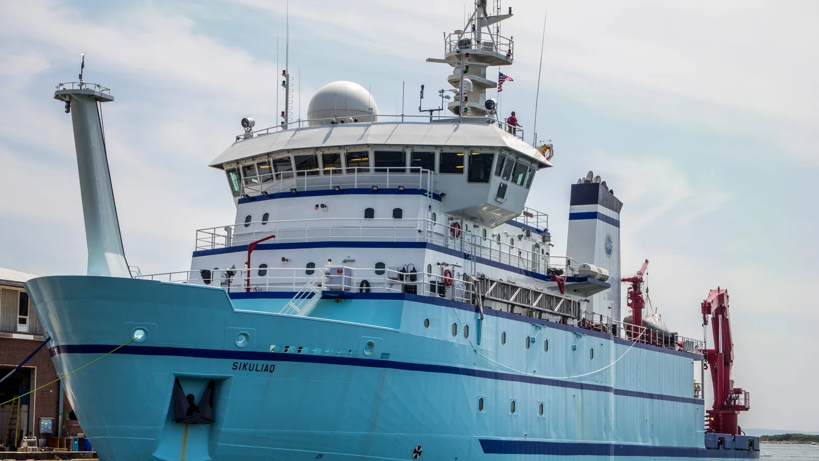 The blue-and-white research vessel Sikuliaq sits at a dock under a pale sky.