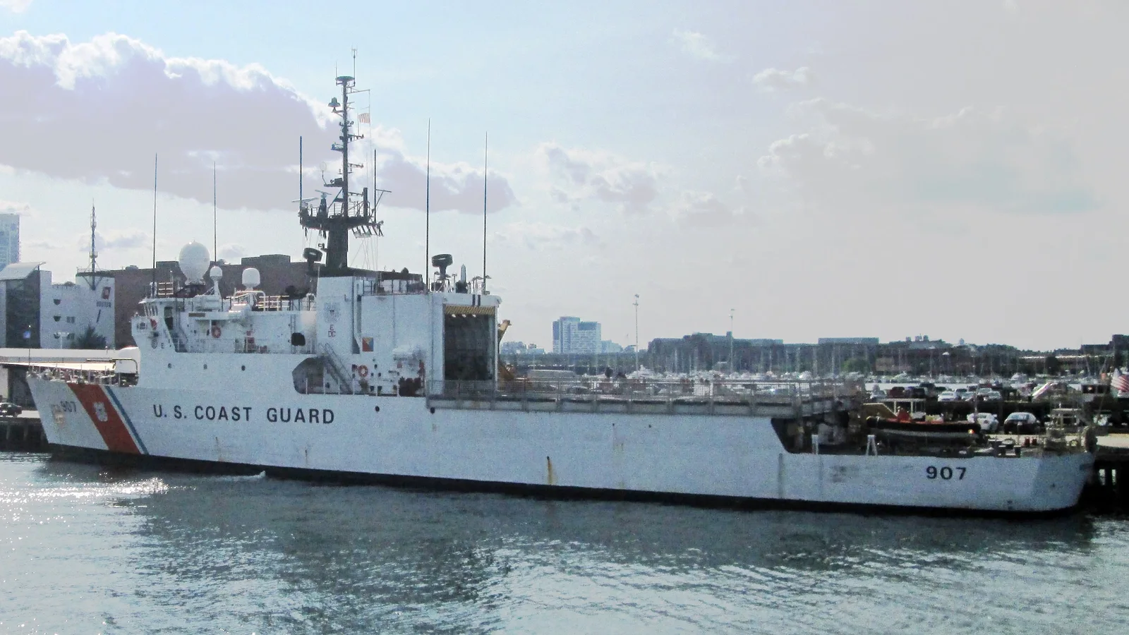 A red-and-white Coast Guard cutter is moored beside brick buildings at a waterfront base.