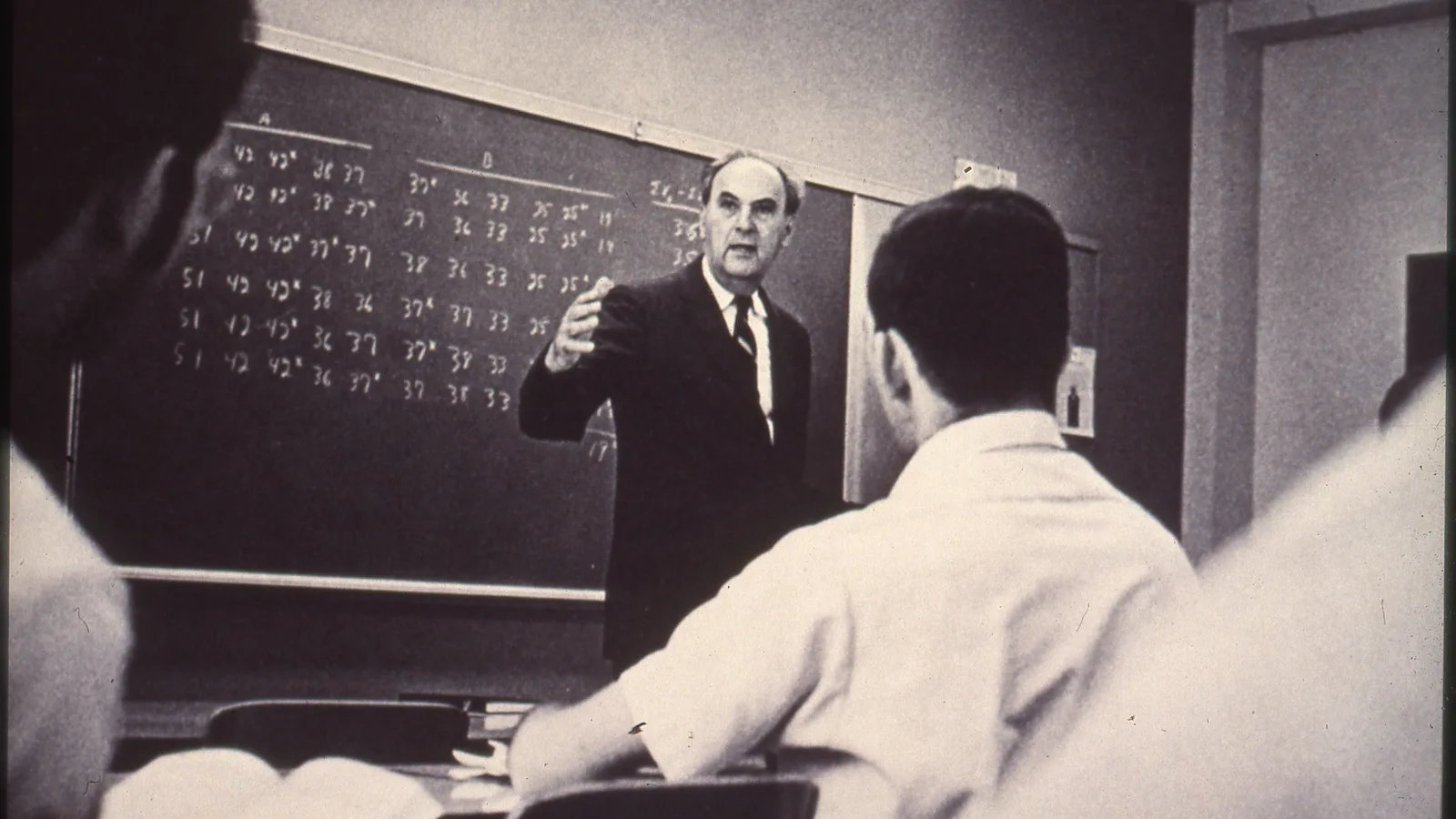 Students sit at desks in a college classroom while an instructor stands at the front.