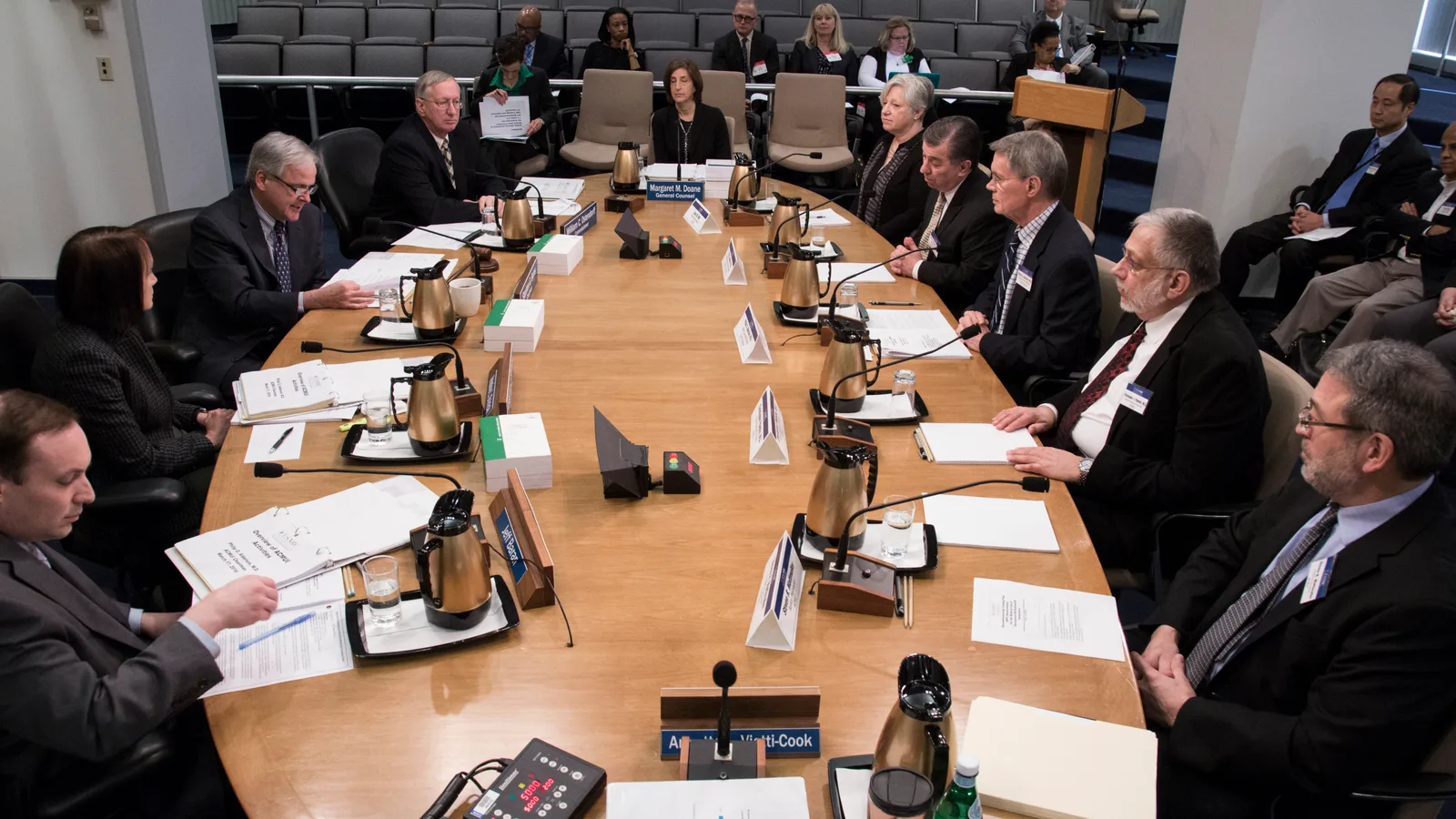 Nuclear Regulatory Commission members and staff sit at a long table during a public meeting.
