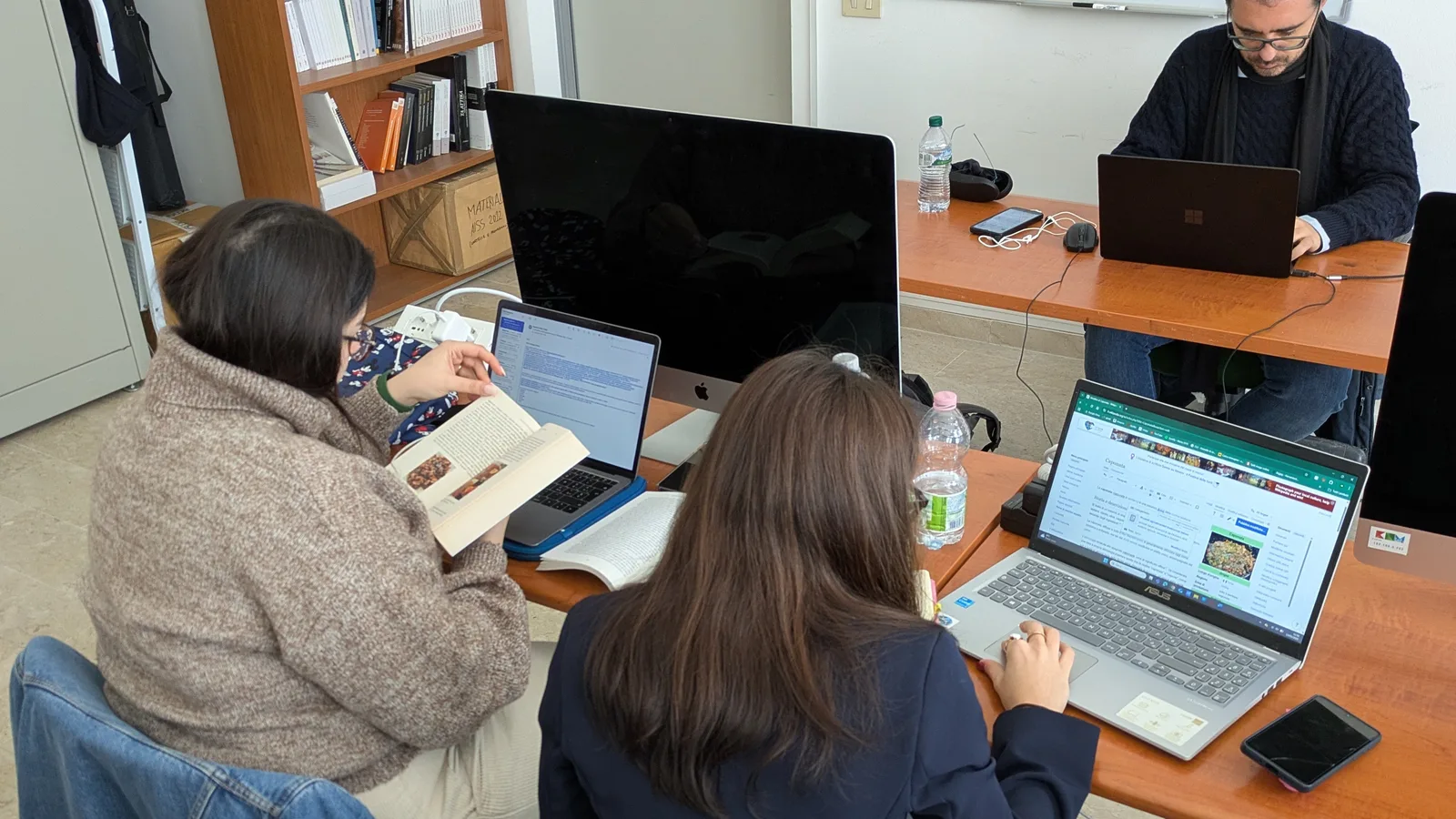Adults working together at laptops around tables during a university hackathon workshop.