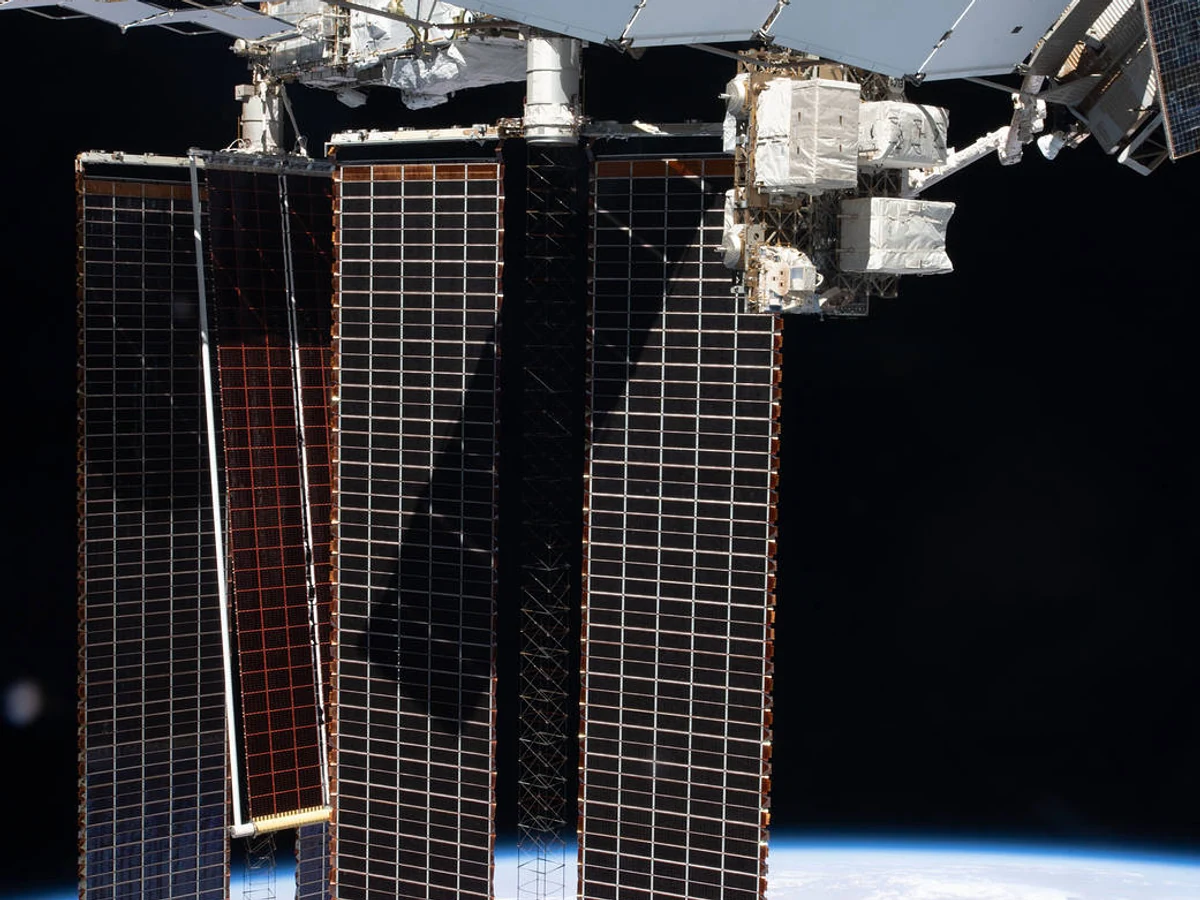 A narrow roll-out solar array deployed in front of the International Space Station's larger legacy solar panels above Earth.