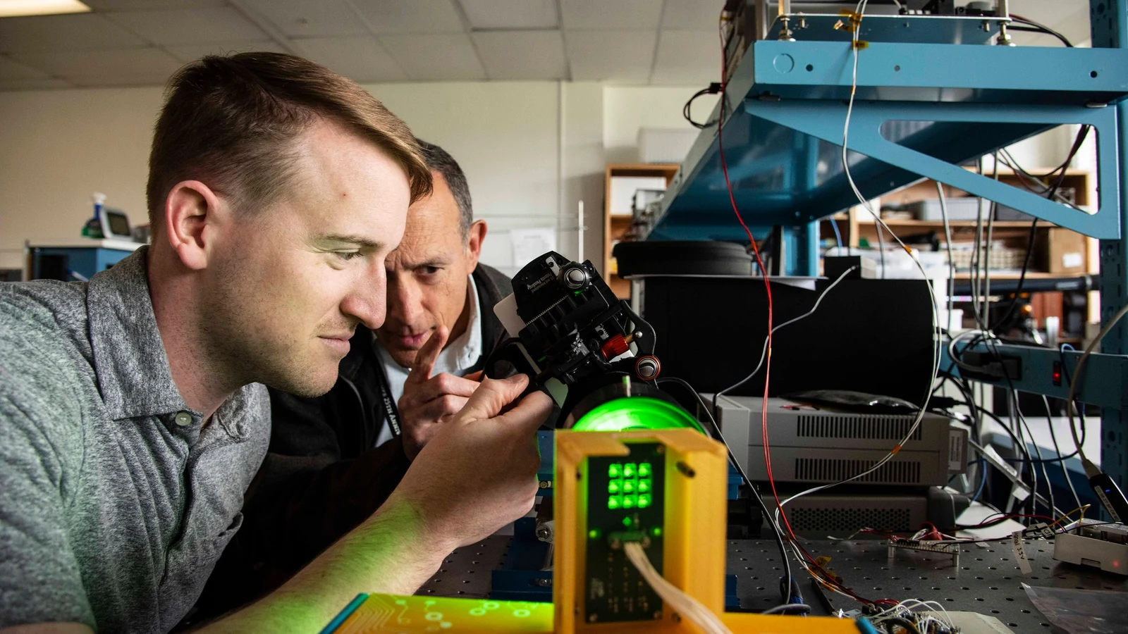 Two engineers inspect a small CubeSat optical-communications payload on a workbench.