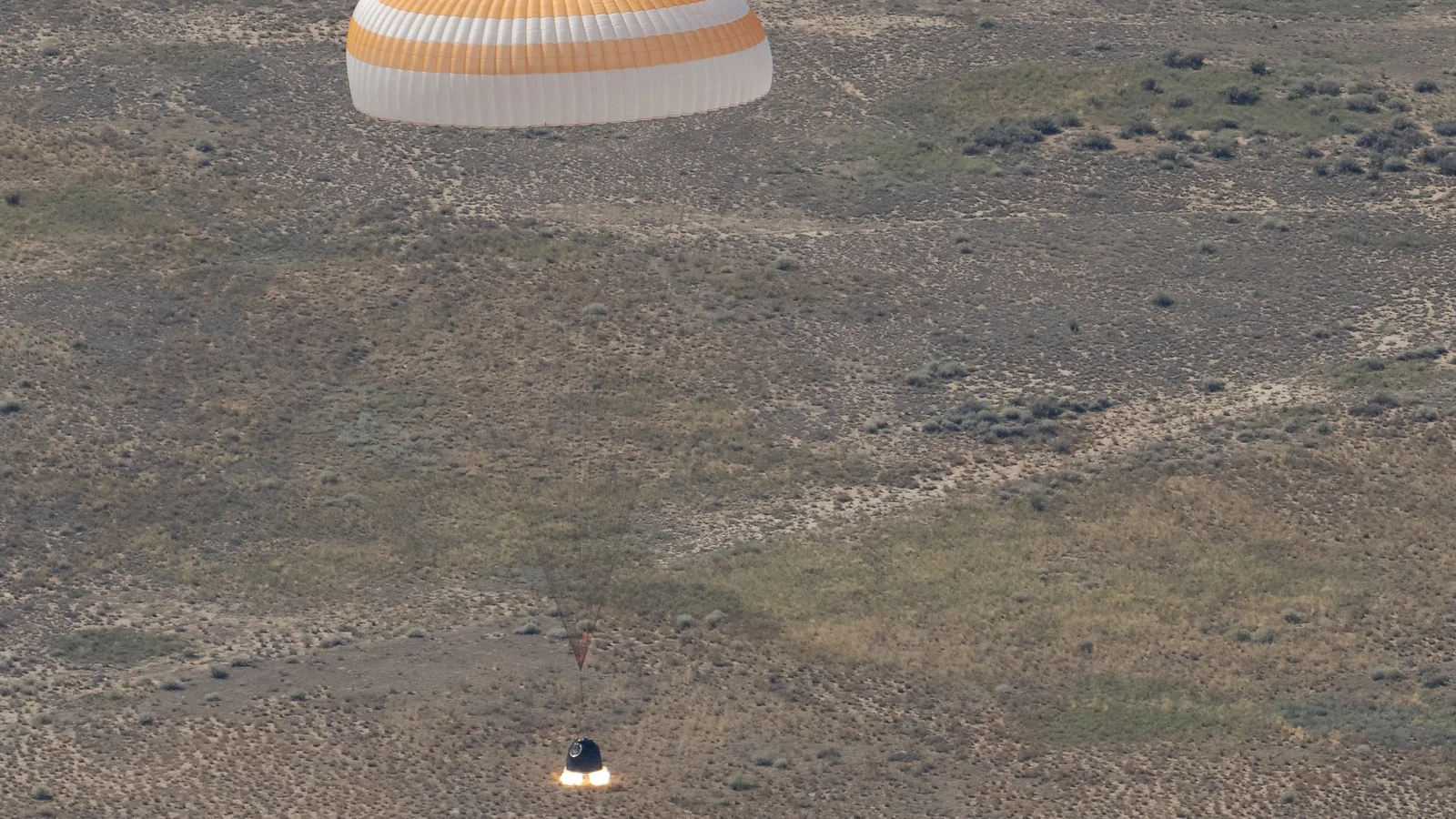 A Soyuz capsule descends under a parachute over the Kazakhstan steppe as recovery vehicles wait below.