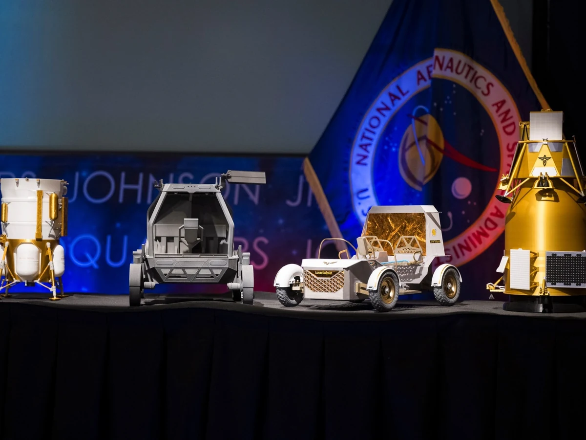 Scale models of two lunar landers and two lunar rovers stand on a stage in front of a NASA flag.