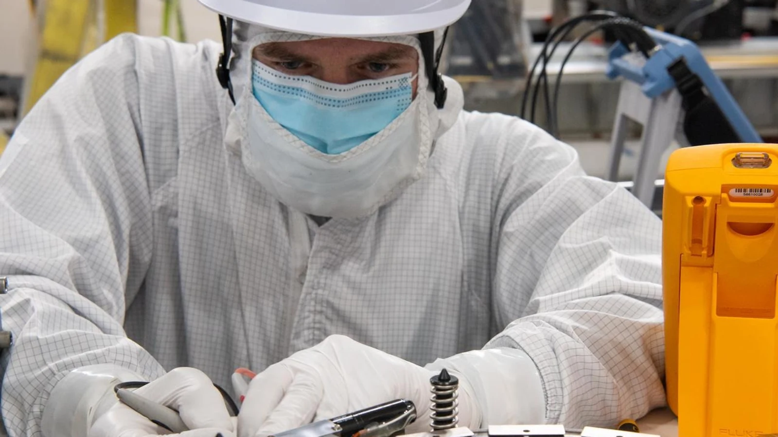 A technician in clean-room clothing works closely on circular spacecraft hardware at a laboratory bench.