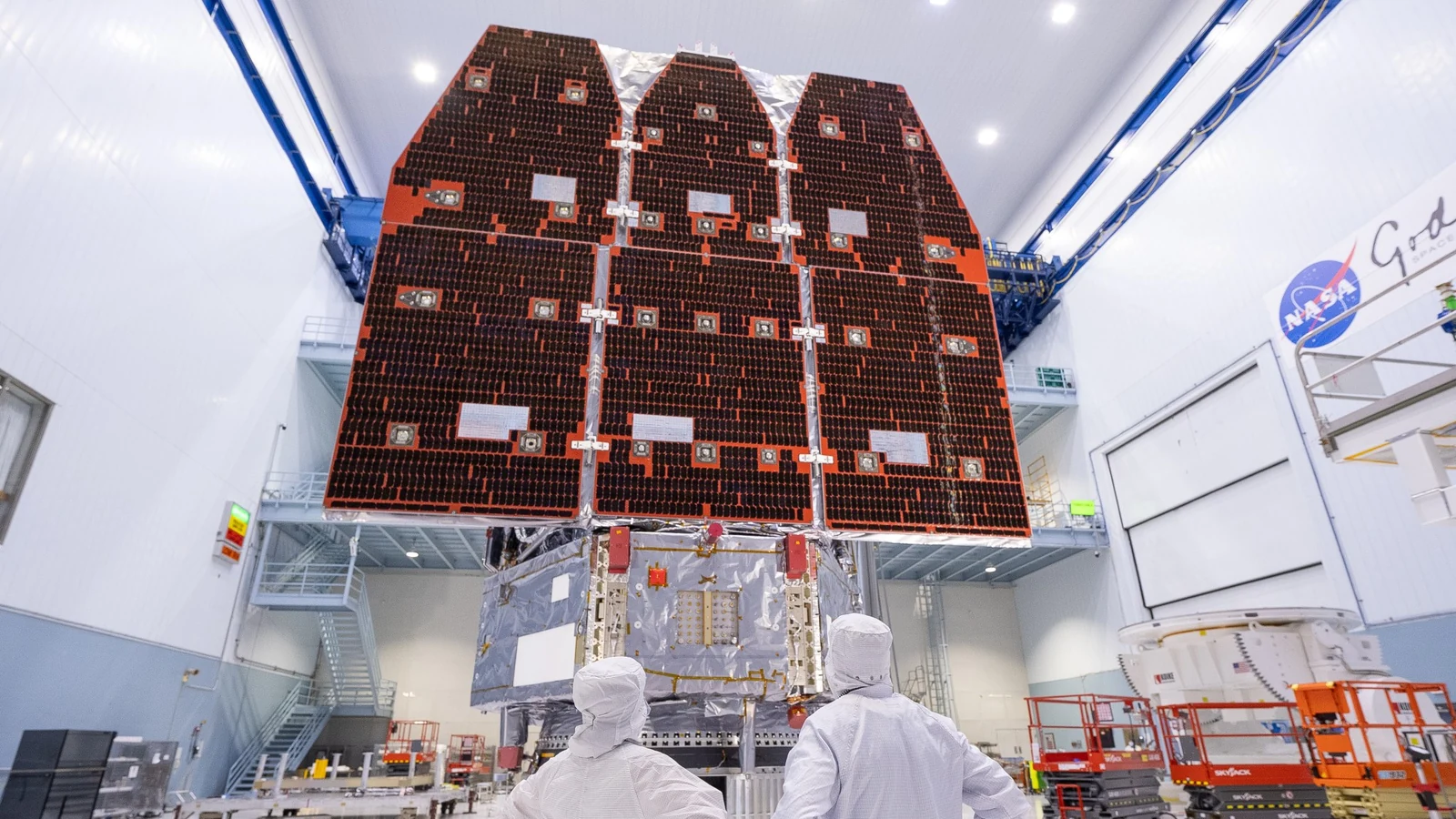 Two technicians in clean-room suits stand beneath the assembled Roman Space Telescope and its deployed solar panels.