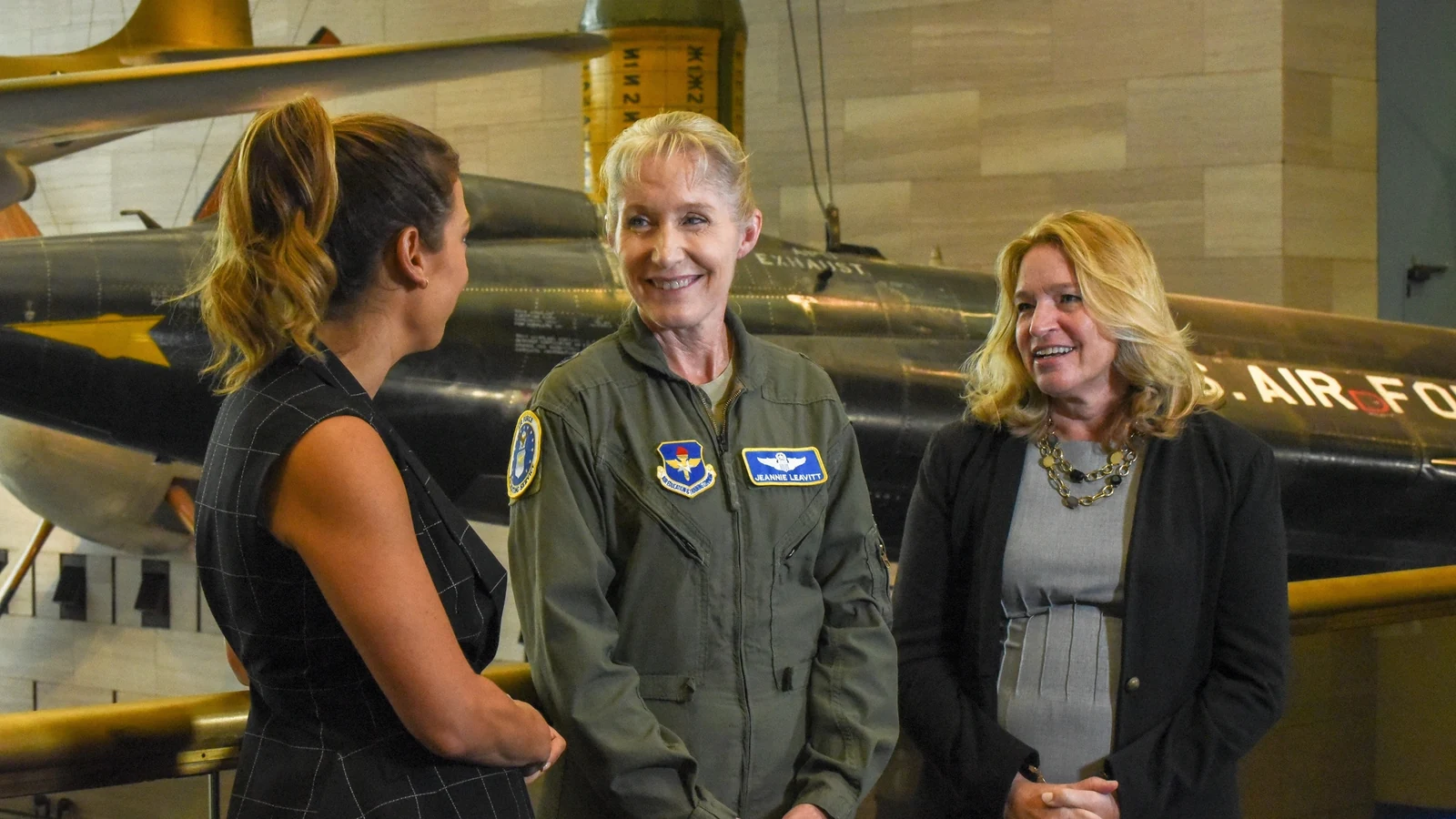 Three women speak in front of a black jet displayed inside the National Air and Space Museum.