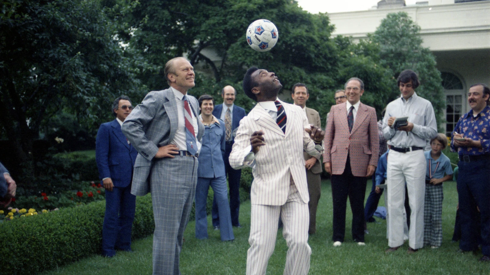 President Gerald Ford looks upward beside Pelé as a soccer ball hangs above them in the White House Rose Garden.