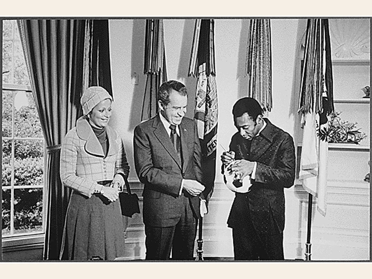 Pelé signs a soccer ball while President Richard Nixon and First Lady Pat Nixon watch at the White House.