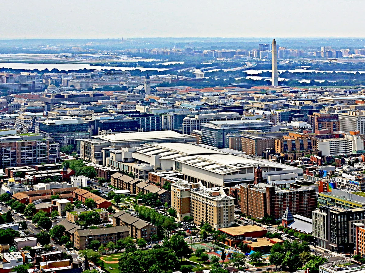 An aerial photograph shows the long white roof of the Walter E. Washington Convention Center amid downtown Washington, with the Washington Monument beyond.