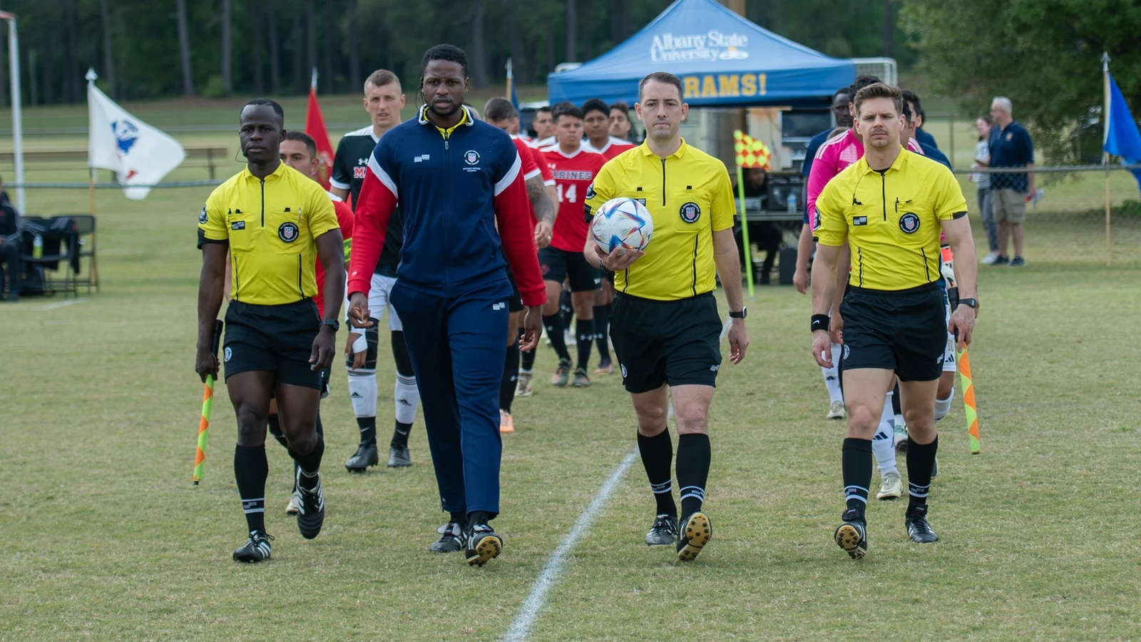Three soccer referees in yellow shirts walk onto a field with team captains before a match.