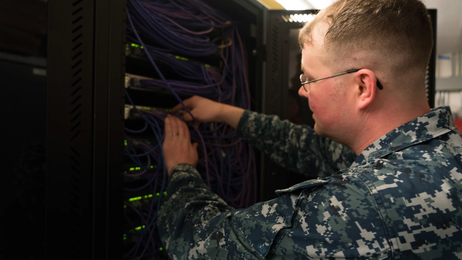 A network specialist works at a desk surrounded by monitors and communications equipment.