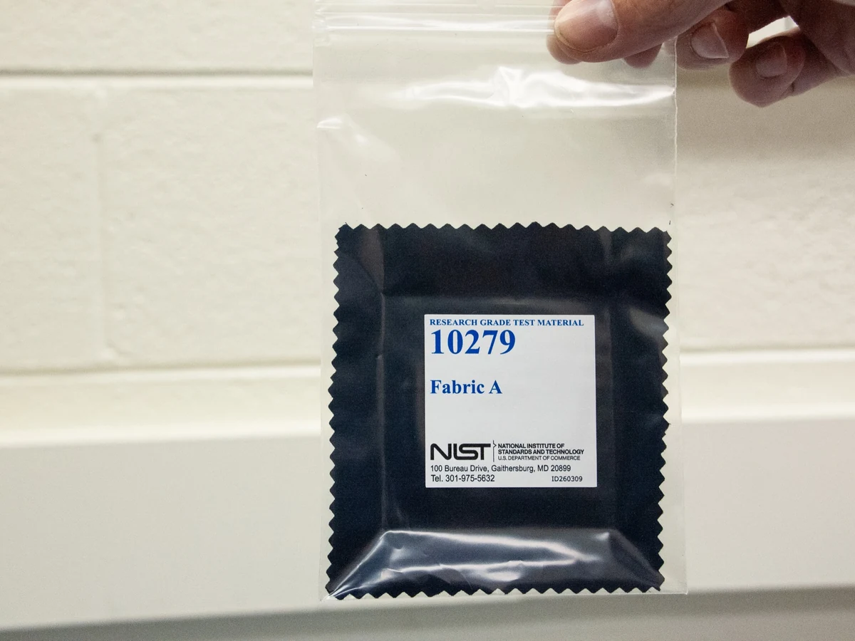 A hand holds a sealed package containing a dark fabric square labeled Research Grade Test Material 10279 Fabric A.