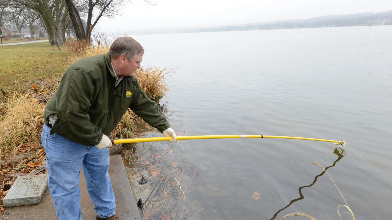 An environmental specialist reaches a long yellow sampling pole into the Mississippi River from the bank.