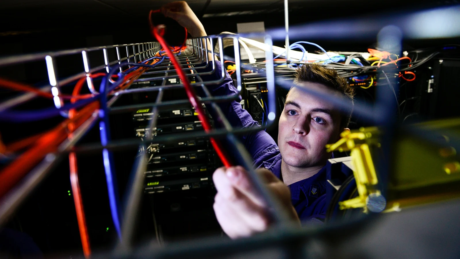 A technician stands between tall server racks and adjusts bundled network cables.