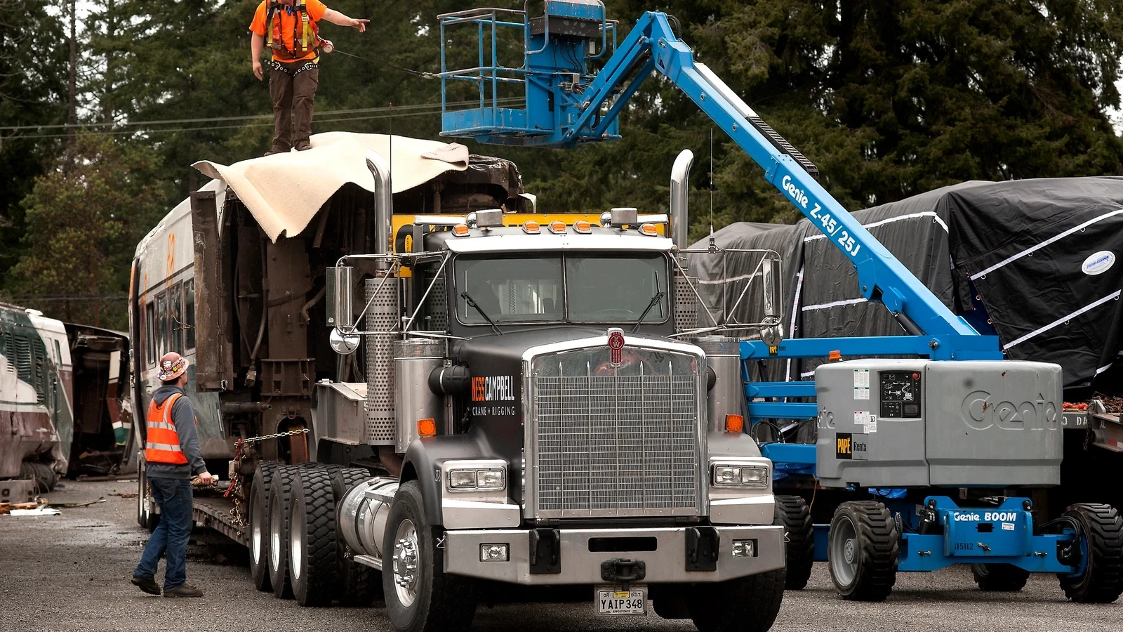 Workers use a lift beside a damaged Amtrak railcar loaded on a truck at Joint Base Lewis-McChord.